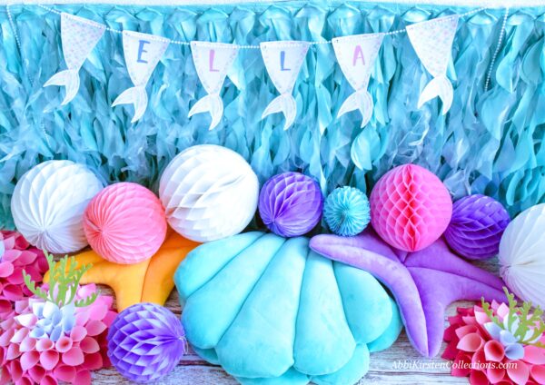 Decorations include a wavy table skirt, with a mermaid tail banner, cloth clam chairs and white, purple, pink and blue honeycomb balls.