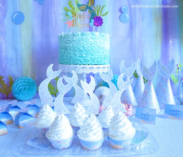 A party spread fit for a Mermaid princess! A platter of cupcakes with aqua blue icing and mermaid tail cupcake toppers, Mermaid party hats, and a gorgeous aqua blue birthday cake.