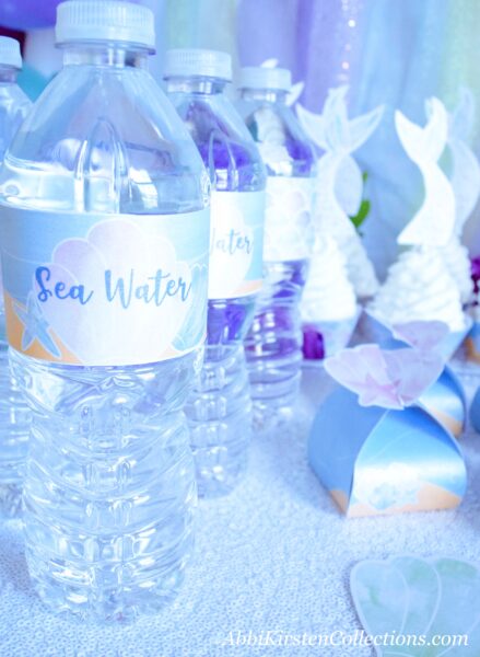 No mermaid party is complete without customized “sea water” water bottle labels. Mermaid tail cupcakes and mini party boxes sit next to bottles of water.