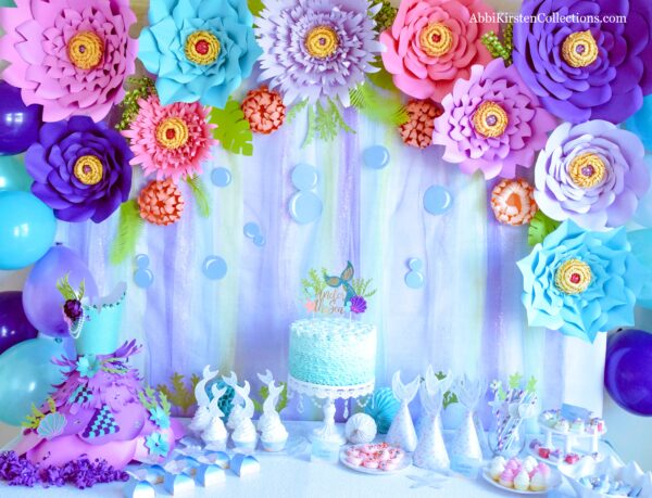 Our Under the Sea Mermaid party backdrop has blue, purple, and green iridescent tones, large purple and blue paper flowers, and paper bubbles.
