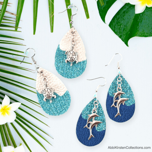 Two sets of faux leather earrings, one white and turquoise with sea turtle charms and the other dark blue and turquoise with dolphin charms, laying next to tropical leaves and flowers. 
 