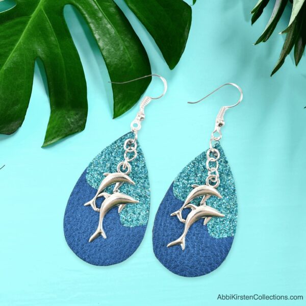 A close up of turquoise and blue faux leather earrings with silver dolphin charms laying on a light blue table surrounded by tropical leaves. 