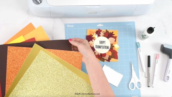 A hand holding fall colored glitter and foil cardstock near a Cricut machine and tools. 