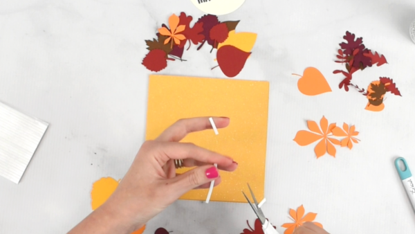 Hands cutting white foam strip tape above supplies for a Thanksgiving Card.  