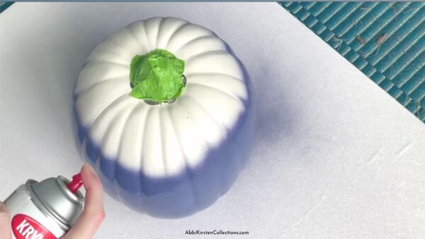 A white craft pumpkin in the process of being spray painted a rich navy blue color. The stem of the pumpkin is wrapped with green painter's tape.