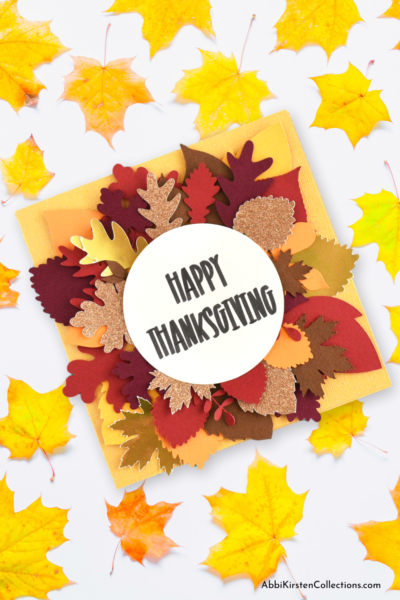 A yellow Happy Thanksgiving Card decorated with a variety of fall leaves sitting on a white table with yellow leaves. 