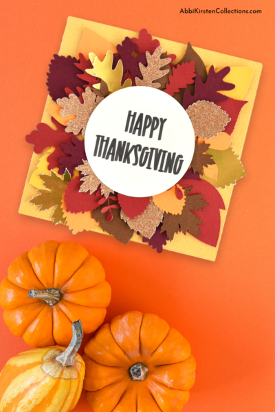 A yellow card with fall leaves and text that reads "Happy Thanksgiving" on an orange table with gourds nearby. 
