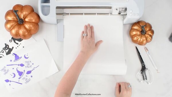 A woman loads a sheet of white vinyl into a Cricut sitting on a white table. Small craft pumpkins, Halloween cutouts, and scissors sit on the table too.