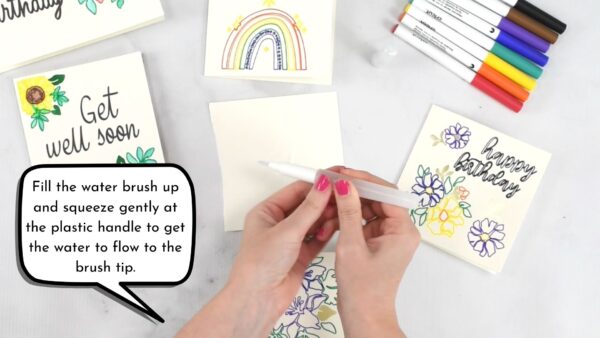 An overhead view of Abbi's hands holding a watercolor brush. Below are half-done printed greeting cards created with a Cricut. A speech bubble says "Fill the water brush up and squeeze gently at the plastic handle to get the water to flow to the brush tip."
