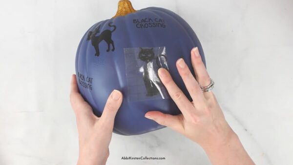 A woman applies a black cat decal to a navy blue painted pumpkin using transfer tape. The pumpkin already has multiple other vinyl cut outs applied around the surface.