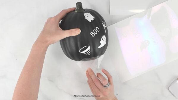 A woman applies white vinyl decals to a small black painted craft pumpkin. The pumpkin has little ghosts and the word "boo" applied all over the surface.