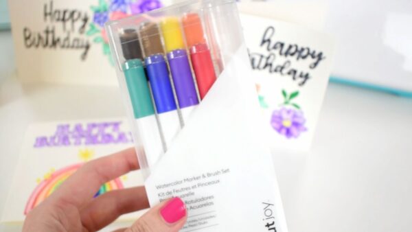 A new Cricut watercolor markers and water brush set is held by Abbi's hand for a close-up. Other cards are blurred in the distance. 