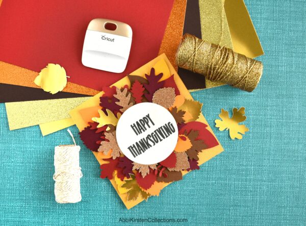 A yellow card decorated with fall leaves and text that reads "Happy Thanksgiving" on top of a turquoise workstation with card supplies nearby. 