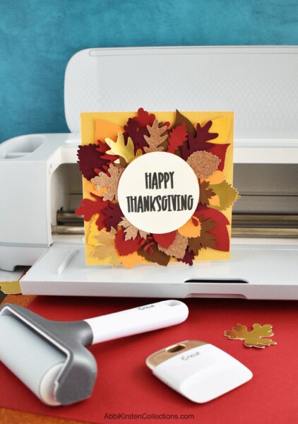A yellow greeting card decorated with fall leaves and text that reads "Happy Thanksgiving" set on a white Cricut machine with Cricut tools nearby. 