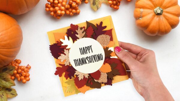 A hand holding a yellow greeting card decorated with fall leaves and text that reads "Happy Thanksgiving". There are orange pumpkins and berries on a white table nearby. 