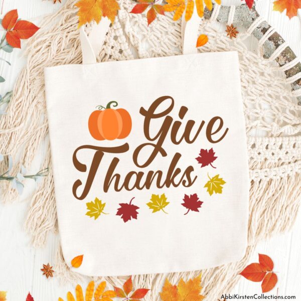 An off-white knitted blanket lays on a white wooden surface with fall leaves at the bottom. On top of the blanket is a canvas tote bag, customized with a Cricut machine and vinyl that says "Give thanks," and is has a pumpkin and autumn leaf embellishments.
