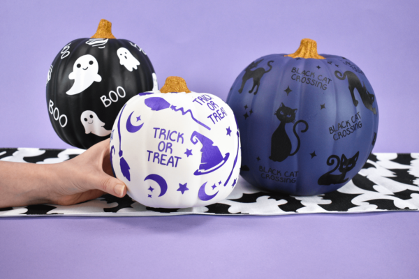 Three decorative pumpkins, painted black, white, and navy blue, covered with Halloween vinyl cutout designs like ghosts, witches hats and brooms, and black cats.