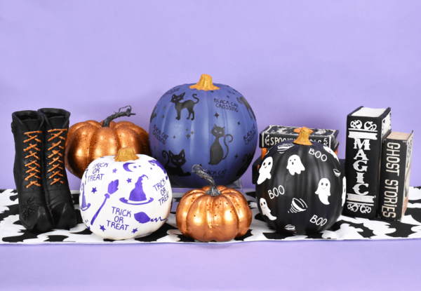 Painted pumpkins decorated with Halloween designs cut from vinyl, set up among festive Halloween decorations. A white, purple, and black pumpkin sit next to a pair of witches boots, spell books, and shiny orange decorative pumpkins.