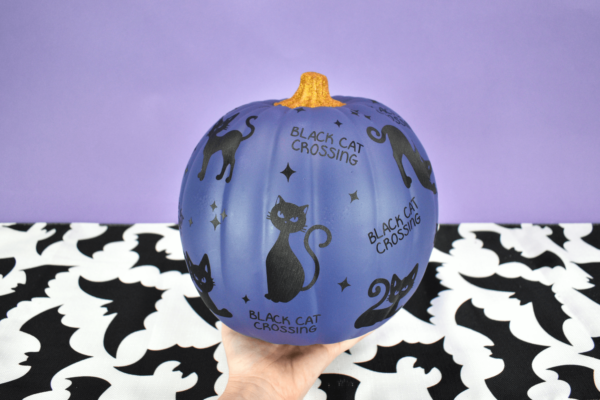 A woman holds a navy blue painted pumpkin in her hands. it's covered with black cat vinyl cut outs, little stars, and words that say "black cat crossing"