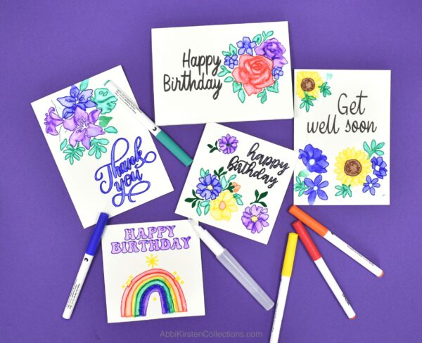 Cricut Watercolor Markers and Brush Set are perfect for paper crafts and DIY cards. These handmade cards say "Happy Birthday," "Get Well Soon," and "Thank You" on a dark blue paper background. Watercolor pens are scattered around. 