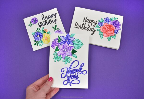 Thank you and happy birthday watercolor cards with Cricut on a dark blue paper background. Each card is white with multicolor watercraft flowers and green leaves. 