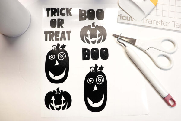 The free trick or treat SVG file for Cricut and Silhouette cutting machines has been made and laid in the center of a white tin and a Cricut weeding tool, detail scissors, and the corner of transfer tape. The finished vinyl has the words "Trick or treat," and two "boos," plus different fun jack-o-lanterns. 
