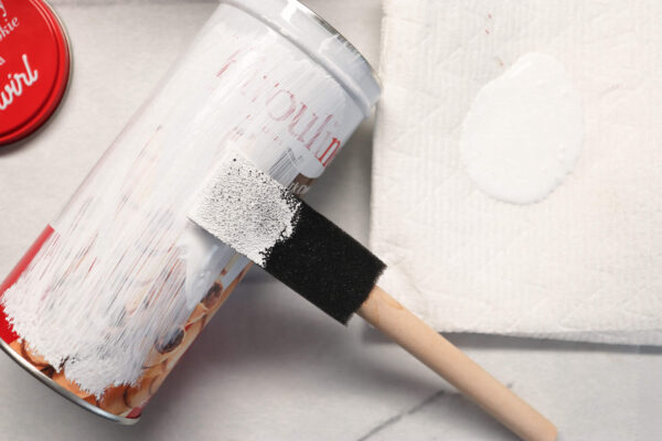 A cookie tin is being painted white with a foam brush. A paper towel holding a puddle of white chalk paint sits to the right of the brush. 