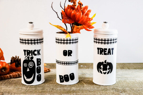 The words "trick or treat," and "boo" are in black vinyl and attached to repurposed tall cookie tins. Fall faux flowers are in the center tin, while the other too have new lids. You can make this Halloween craft using repurposed tins. 