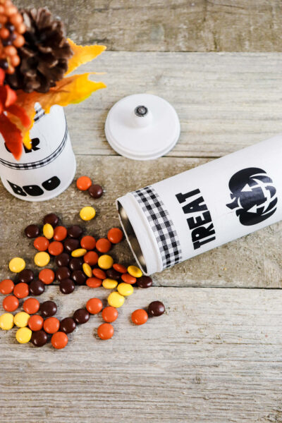 Trick or treat cookie tins decorated with Cricut vinyl and chalk pain are seen from above. One tin is used as a vase for faux fall leaves and pinecones, and another is laying on a table spilling fall-colored candies. 