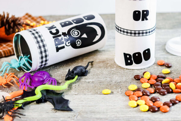 Two repurposed tall cookie tins, one standing and one laying on a table, are white and decorated with images of pumpkins, and the words "trick," "boo," and "or." Colorful fake bugs and fall-covered candies are on the table as well. 