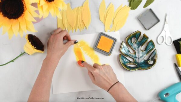 Amid finished crepe paper sunflowers and cut-out petals and supplies, Abbi Kirsten's hands add distress ink to the yellow crepe paper petals. 