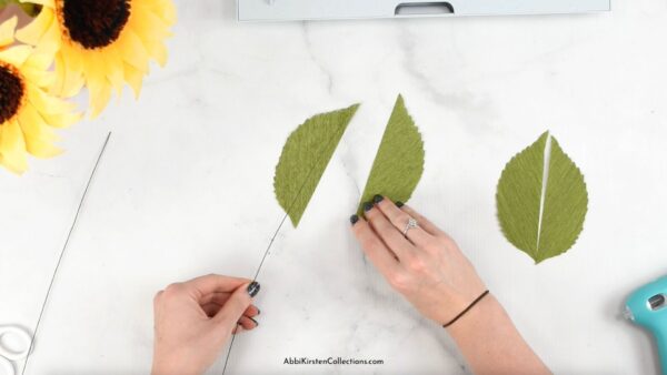 Two crepe paper sunflowers dot the top of the table, while Abbi Kirsten's hands glue paper green leaves to floral wire. 