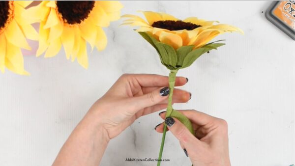 Abbi Kirsten's hand gently hold the stem of a crepe paper sunflowers while adding paper leaves. 