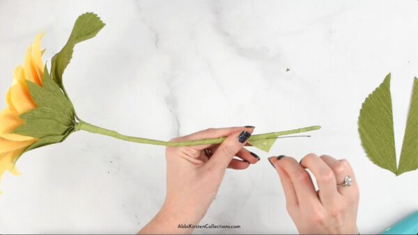 Overhead view of Abbi Kirsten's hands assembling the stem and leaves of a crepe paper sunflower. 