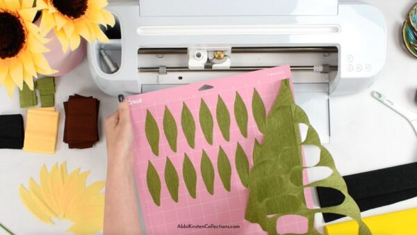 Abbi Kirsten demonstrates how to peel the crepe paper off a Cricut fabric mat. Below is a cutting machine and cut-outs for the sunflower paper flower craft. 