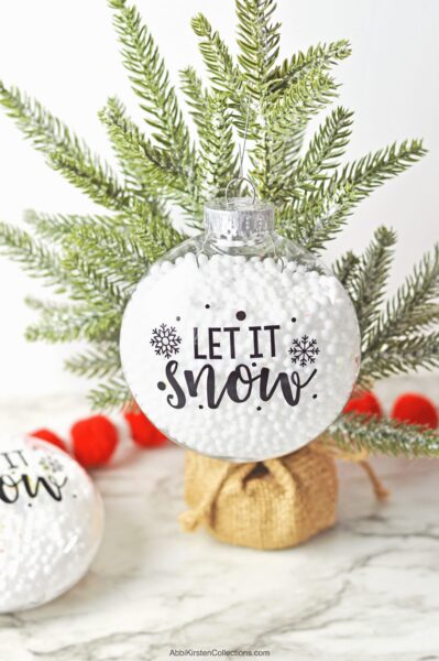 A round, fillable Christmas ornament hangs on a small pine tree on a marble tabletop. Part of a similar decoration lies nearby. The bauble has vinyl text that reads "Let it Snow."