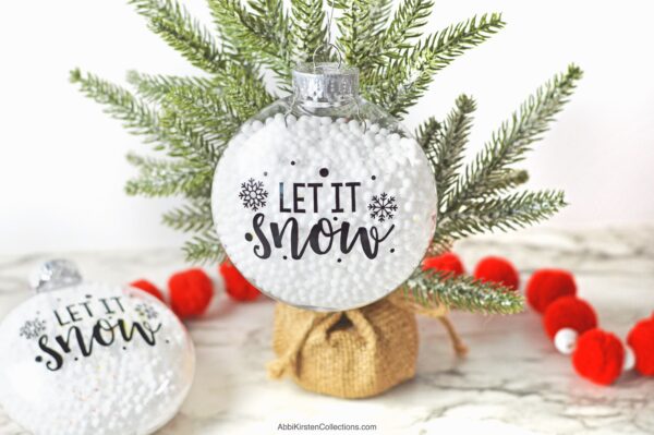  A tiny tabletop Christmas tree holds a clear, fillable Christmas ornament filled with white snowballs and text that reads "Let it Snow." A similar decoration lays next to the tree and a red-ball garland.