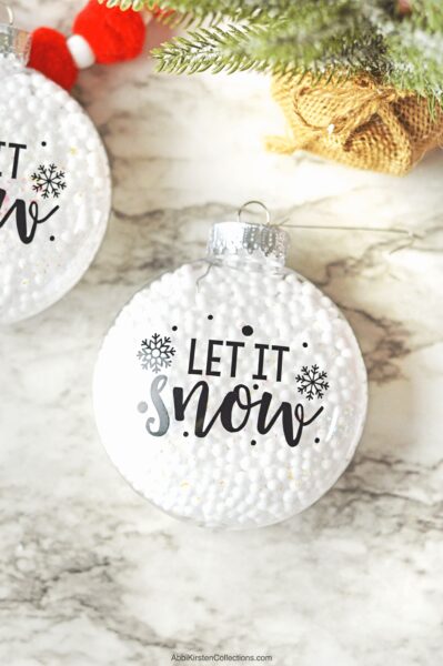 A fillable Christmas ornament made of clear plastic and full of white snowballs lays on a festive tabletop next to a similar decoration, only half visible. The text on the ornaments says, "Let it Snow."