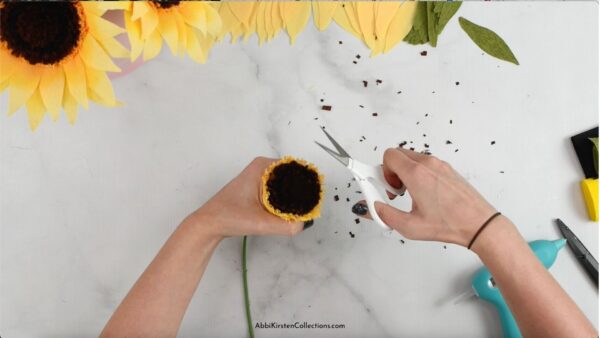 Abbi Kirsten's hands hold the crepe paper sunflower center while using detail scissors to trim the brown center. Bits of brown crepe paper freckle the work table. 