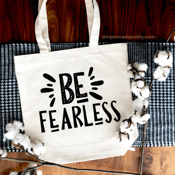 A beige canvas tote lays on a black and white blanket decorated with cotton stalks. The tote says "Be fearless" in a black typography SVG design.