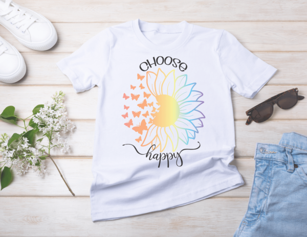 Tennis shoes, jeans, sunglasses, and flowers lay on white wood with a white t-shirt in the middle. The t-shirt has a rainbow-colored sunflower with the left side turning into butterflies. The black text on the shirt says, "Choose Happy."  