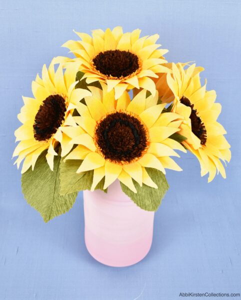 A finished bouquet of crepe paper sunflowers with paper greenery sit in a light pink vase against a blue  background. 