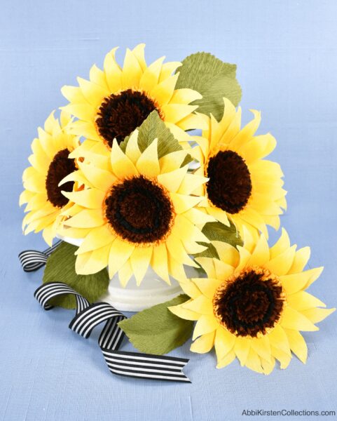 A bouquet of bright paper sunflowers in a white vase with decorative black and white striped ribbon. 