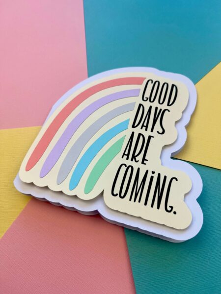 A rainbow-shaped card sits on the center of a spread of varying colors of cardstock. There is a modified rainbow next to the words, "Good days are coming." The design is available as a  free SVG design. 