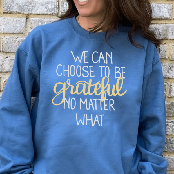 A close-up of a woman wearing a blue sweatshirt while standing in front of a light brick wall. The sweatshirt's text reads, "We can choose to be grateful no matter what." This design is available as a free cut file.