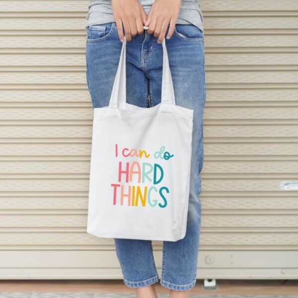 The "I can do hard things" free cut file was used to create the rainbow text on the beige totebag held by a woman against a garage door.  