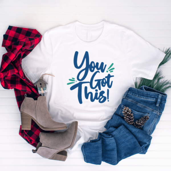 Gray shoes, a red and black checkered flannel shirt, jeans, and pine cones surround the white t-shirt in the center, with a text graphic on the front that reads, "You Got This!"