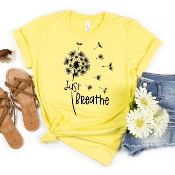 A yellow t-shirt with a dandelion blowing in the wind with the words "Just Breathe" underneath it. Sandals and folded jeans with daisies on them lay next to the shirt. 