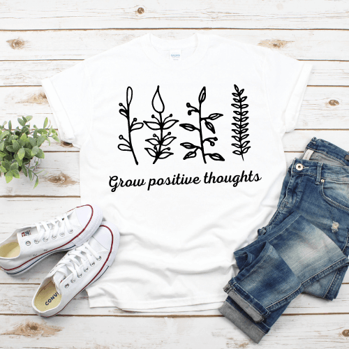 A white t-shirt lies on a white wooden background between jeans and white tennis shoes. The shirt has outlines of leaf stalks and text that says, "Grow positive thoughts."