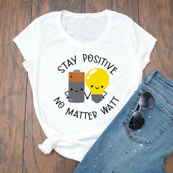 A white t-shirt on a wooden floor, with jeans and sunglasses to the right. The t-shirt has a battery and a lightbulb, smiling and holding hands with the text "Stay Positive No Matter Watt."